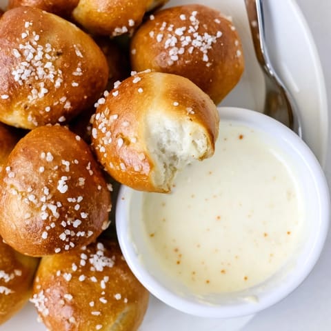 Freshly baked Pretzel Bites with Cheese Dip rest on a wooden board next to a steaming bowl of golden sauce.