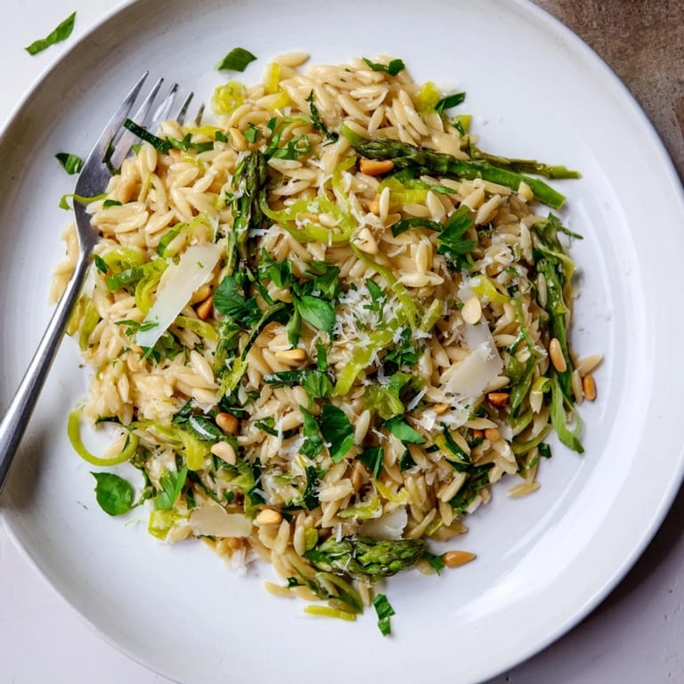 A close-up view of the Asparagus Lemon Orzo Salad, featuring delicate green asparagus shavings, golden toasted pine nuts, and grated Parmesan cheese glistening with a zesty lemon vinaigrette.